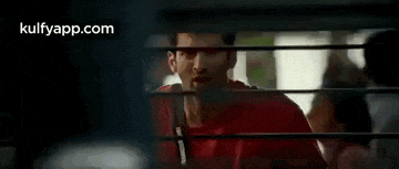 a man in a red shirt is standing in front of a window behind bars .