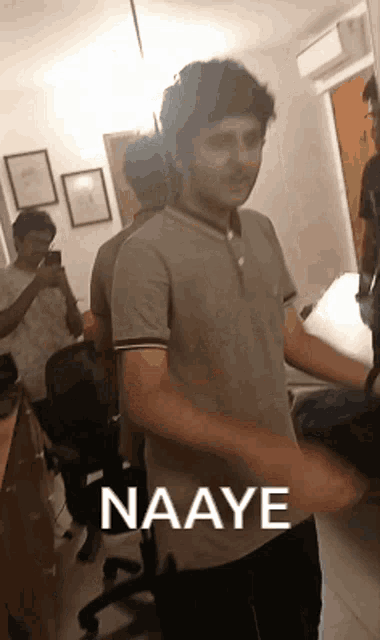 a man in a gray shirt is standing in a room with a group of people and says naaye .