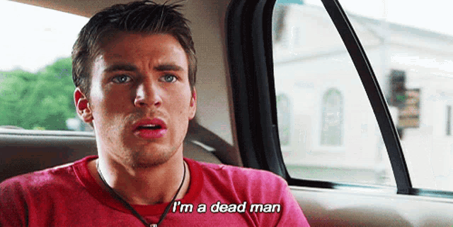 a man in a red shirt is sitting in a car and says i 'm a dead man