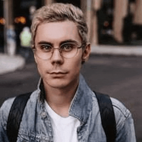 a young man wearing glasses and a denim jacket is standing in front of a building .