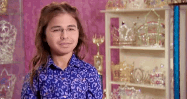 a young boy wearing glasses and a blue shirt is standing in front of a shelf full of tiaras .