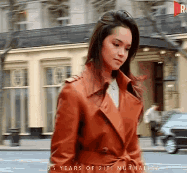 a woman wearing an orange trench coat is walking down a city street