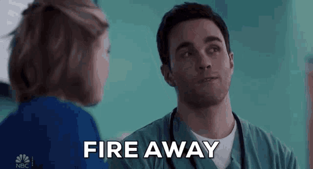 a man in scrubs is talking to a woman in a hospital room and saying fire away .