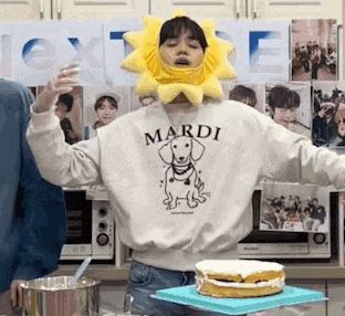 a person wearing a sun hat and a mardi sweatshirt is standing in front of a cake on a table .