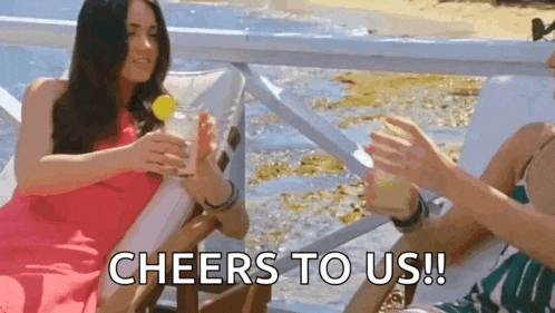 two women are sitting in chairs on the beach holding drinks and a sign that says `` cheers to us ! ''