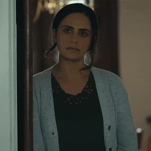 a woman in a black top and a grey cardigan is standing in a doorway looking at the camera .