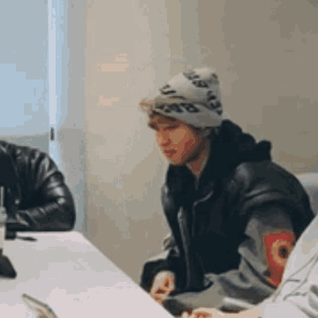 a man wearing a beanie is sitting at a table with a laptop and a cell phone .