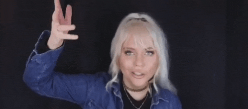 the woman is wearing a blue jacket and making a peace sign with her hands .