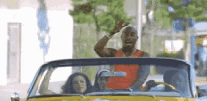 a man in a red shirt is driving a yellow convertible car with two women .