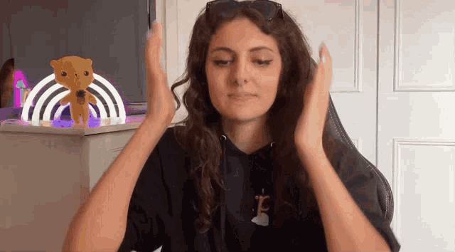 a woman wearing sunglasses and a black hoodie is sitting in a chair with her hands on her head .