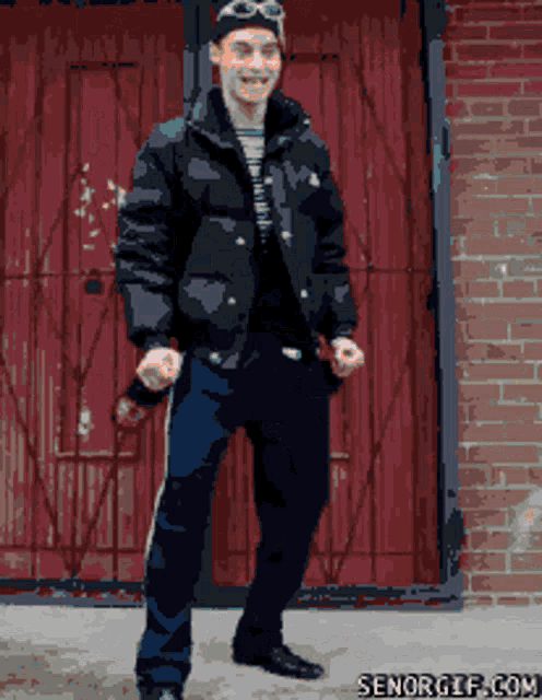 a man in a black jacket stands in front of a red door with senorgif.com written on the bottom