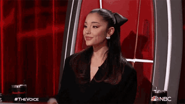 ariana grande is wearing a black jacket and earrings while sitting in front of a red curtain on the voice .