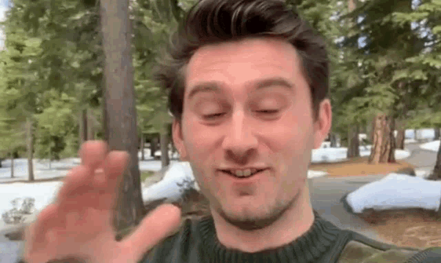 a man in a green sweater is waving his hand in front of a snowy forest .