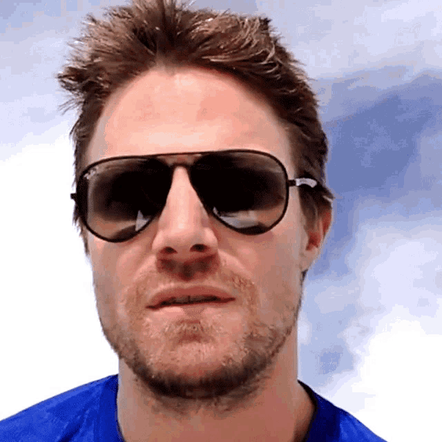 a man wearing ray-ban sunglasses and a blue shirt looks at the camera