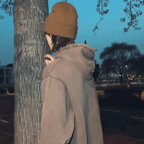 a person wearing a beanie and a hoodie stands next to a tree