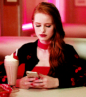 a woman is sitting at a table with a milkshake and looking at her cell phone