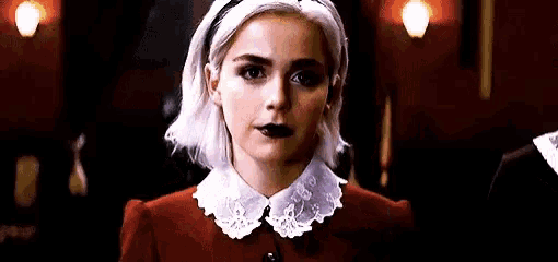 a woman with white hair and black lipstick is wearing a red jacket and white collar .
