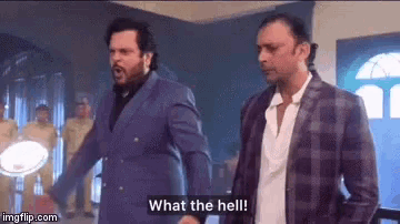 two men in suits are standing next to each other in a room and one of them is saying `` what the hell '' .