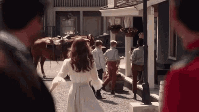 a woman in a white dress is walking in front of a group of people and a horse .