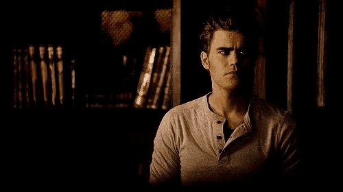 a man in a gray shirt is sitting in front of a bookshelf .