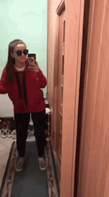 a girl wearing sunglasses and a red jacket is taking a selfie in a hallway