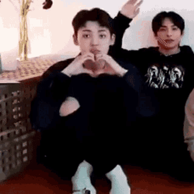 a man is making a heart shape with his hands while sitting on the floor next to another man .