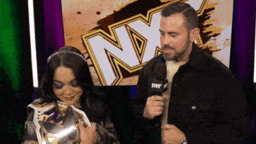 a man is holding a microphone while a woman holds a wrestling championship belt in front of a nxt logo