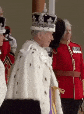 a man wearing a crown and a fur coat is standing in front of a group of people in uniform .