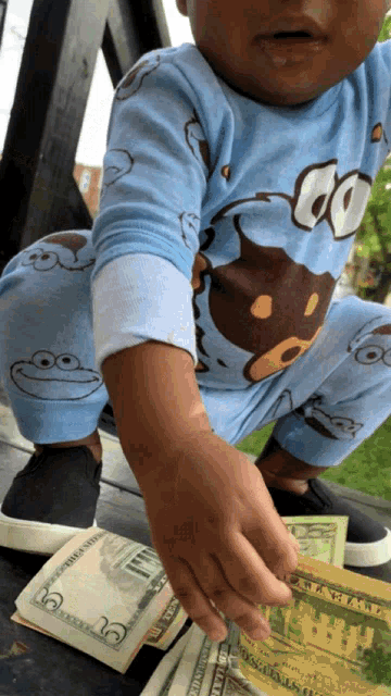 a child wearing a blue monkey pajama is reaching for a stack of 20 dollar bills
