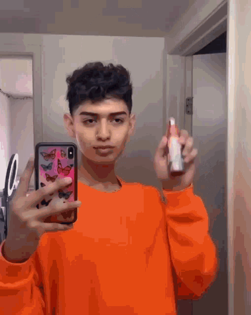 a young man in an orange sweater is taking a selfie with his phone while holding a spray bottle .