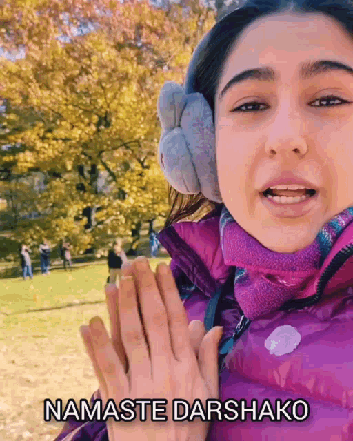 a woman wearing ear muffs and a purple jacket says namaste darshako