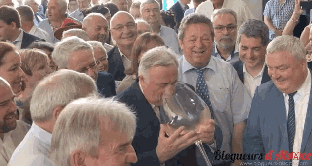 a group of people are gathered around a man holding a wine glass with the words blogeurs d' alsace on the bottom right