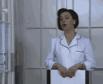 a woman in a white coat is standing in front of a window and holding a folder .
