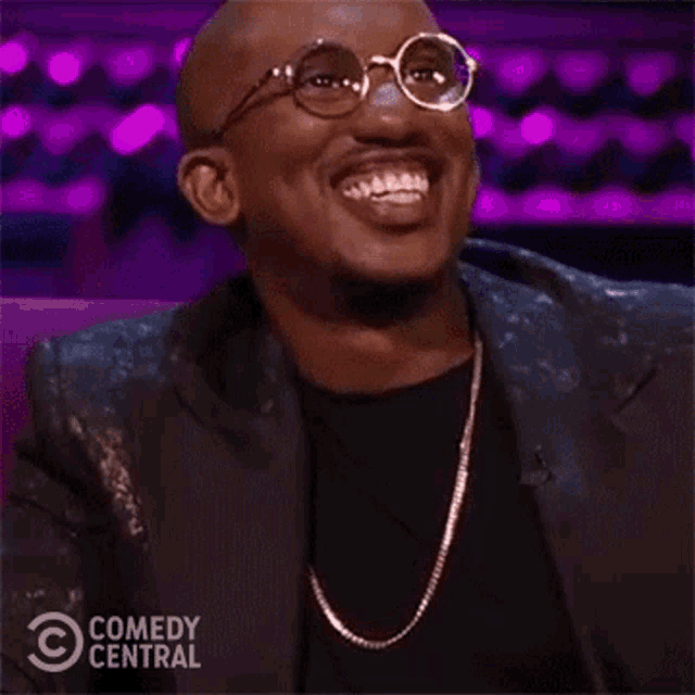 a man wearing glasses and a black jacket is laughing while sitting in a chair .