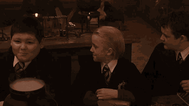 a group of boys are sitting at a table and one of them is wearing a black robe with a crest on it