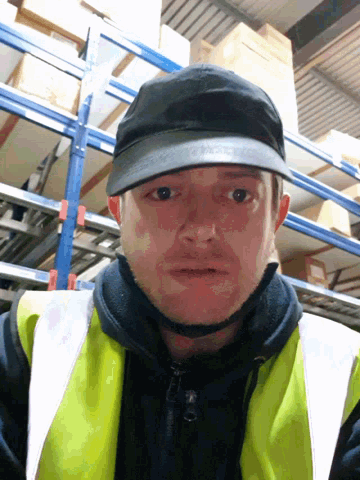 a man wearing a yellow vest and a black hat stands in front of a warehouse