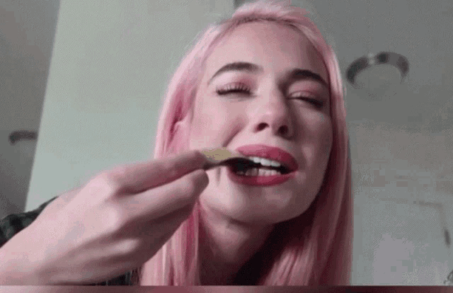 a woman with pink hair is eating something with her eyes closed