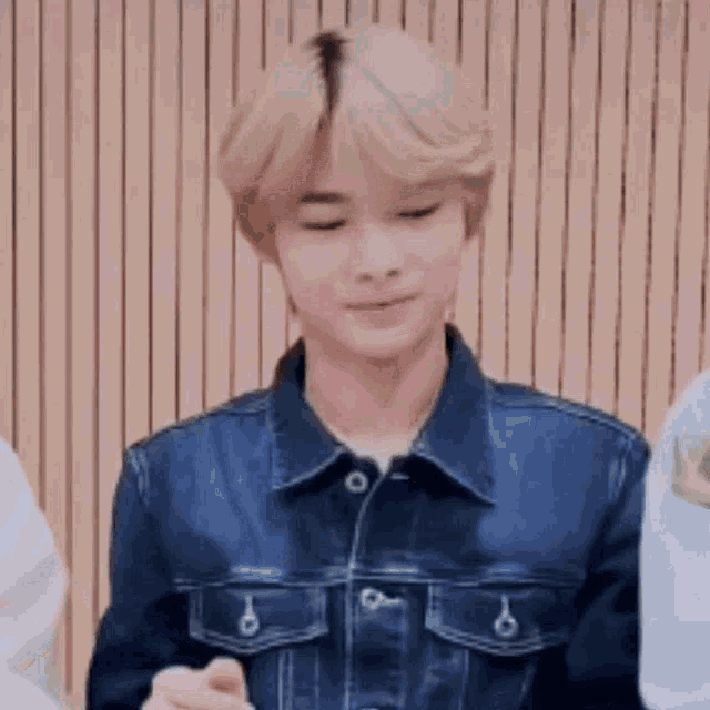 a young man with blonde hair is wearing a denim jacket and standing in front of a wooden wall .