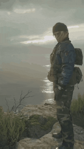 a man in a blue jacket and camo pants stands on a cliff overlooking a body of water