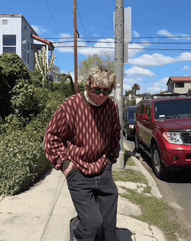 a man wearing sunglasses and a red sweater stands on a sidewalk next to a red suv