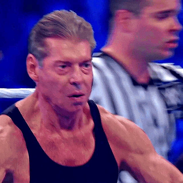 a man in a black tank top stands in a ring with a referee in the background