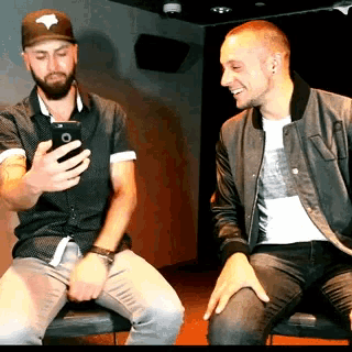 a man taking a selfie with his phone while sitting next to another man