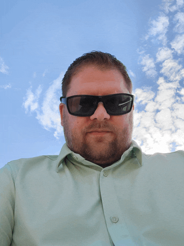 a man wearing sunglasses and a green shirt stands in front of a blue sky with clouds