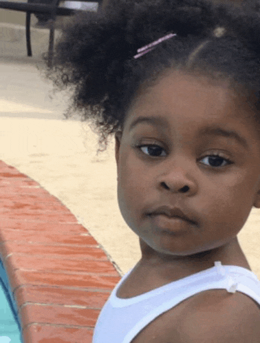 a little girl in a white tank top is looking at the camera