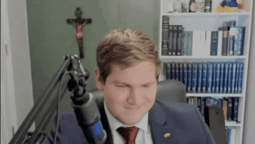 a man in a suit and tie is sitting in front of a microphone in a room with a cross in the background .