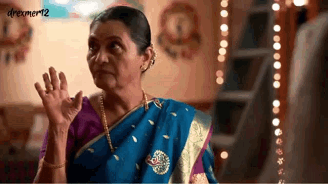 a woman in a blue and purple saree waves her hand in front of a sign that says drexmer12