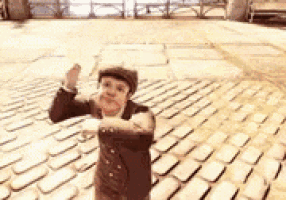 a little boy in a suit and hat is standing on a brick pavement .
