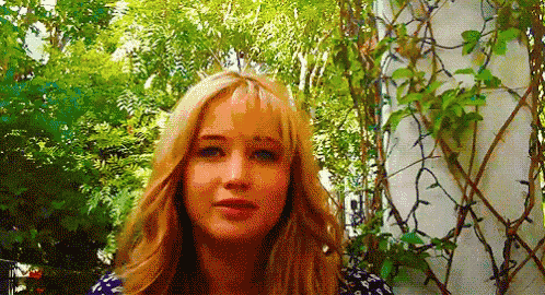 a woman with long blonde hair is standing in front of a lush green forest .