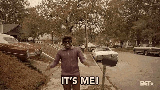 a man in a purple sweater is standing in front of a mailbox and says it 's me .