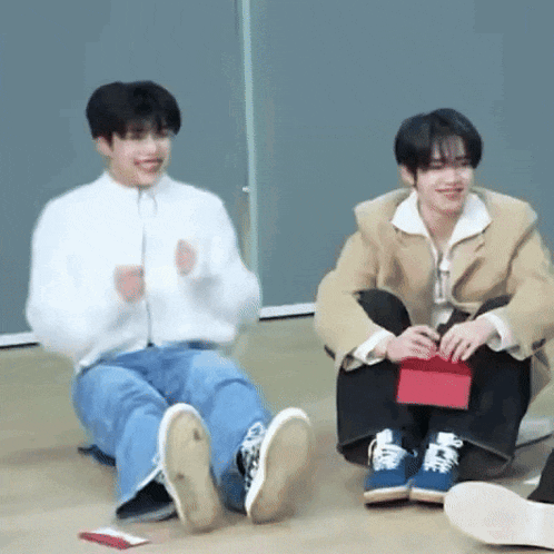 two young men are sitting on the floor with their legs crossed and one of them is holding a red box .